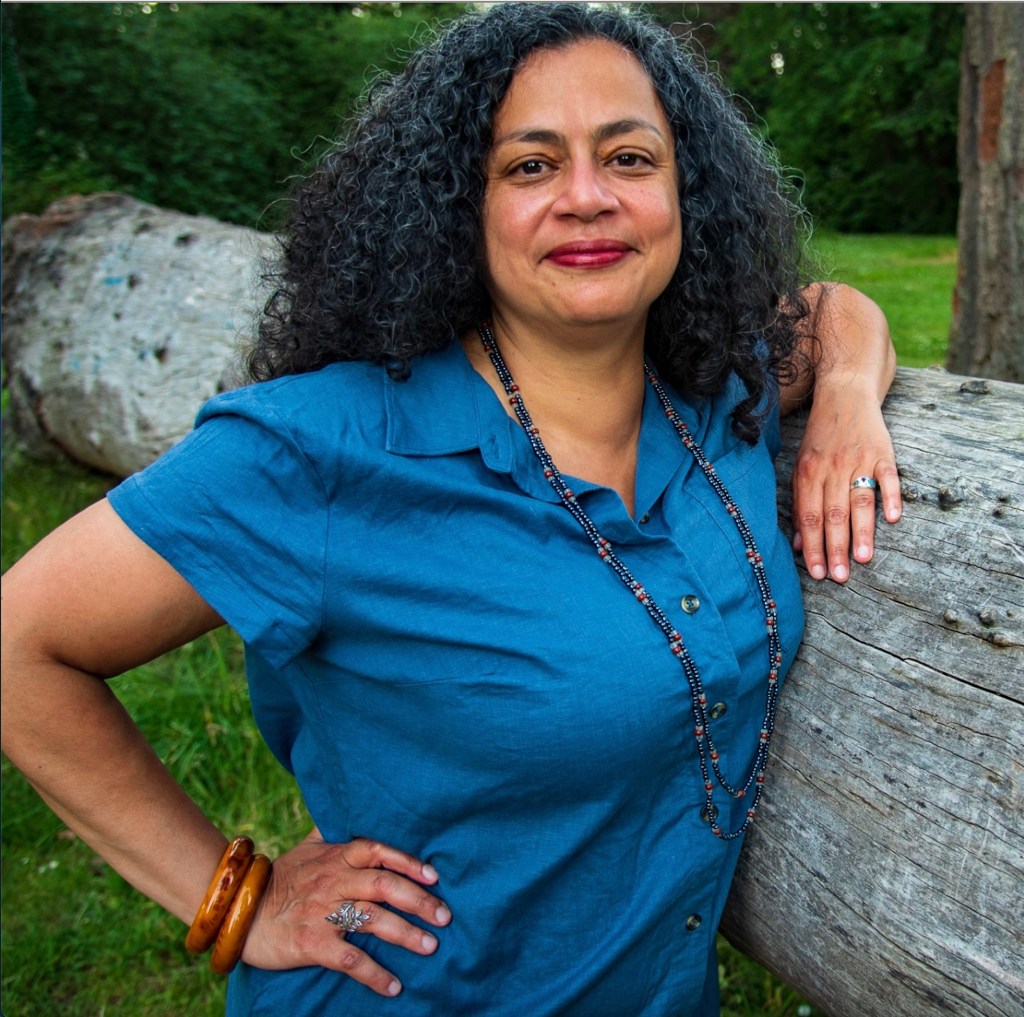This is a headshot of Aisha.  She is wearing a blue short sleeve shirt and leaning up against a tree trunk.