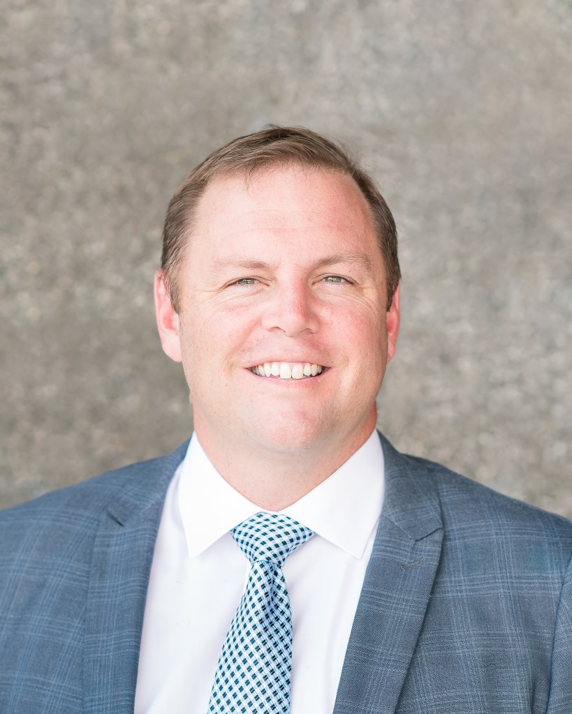 This is a headshot of Dr. Rucks.  He is wearing a blue plaid suit jackets, a white shirt and a light blue polka dotted tie.