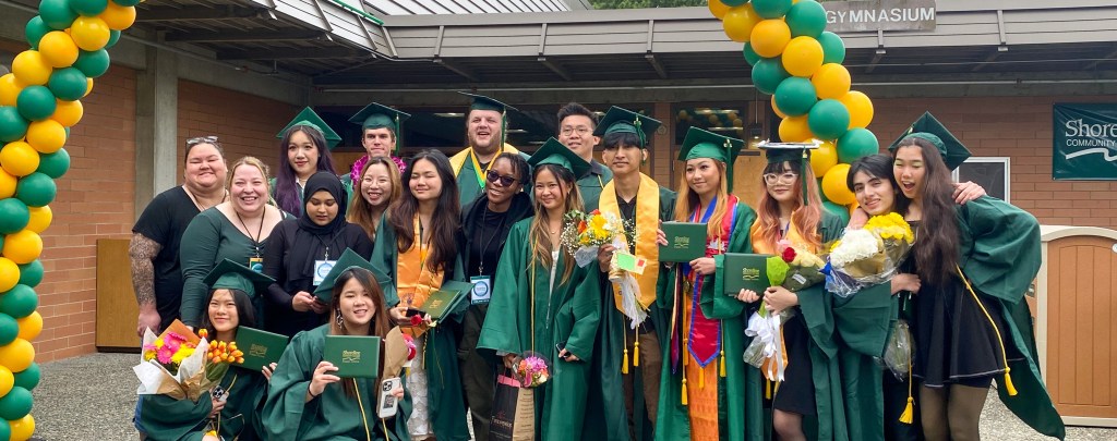 A group of students in graduation regalia gathered under an archway of green and yellow balloons.